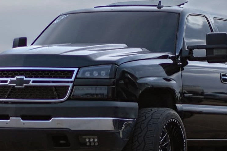 A black lifted Chevrolet pickup truck with large off-road tires and custom wheels, parked outdoors between other vehicles under an overcast sky.
